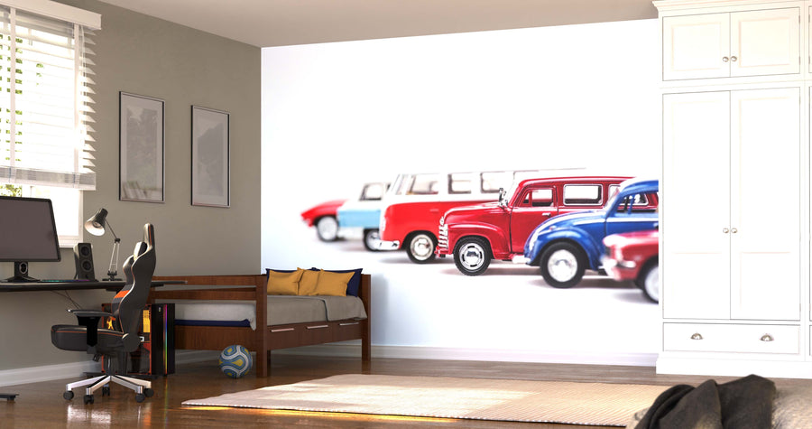 A modern bedroom with a desk, computer, bed, and white wardrobe features the Toy Cars Wall Mural behind the bed. This removable wallpaper showcases vintage red, blue, and white cars, adding an artistic touch as sunlight streams through the blinds.