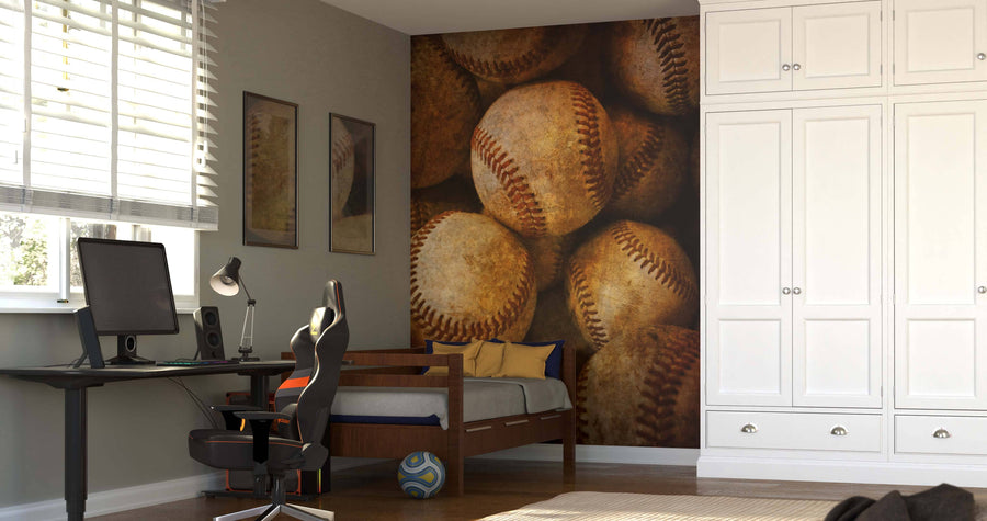 A modern bedroom featuring a Vintage Baseballs Wall Mural, a single bed with blue and yellow pillows, a desk with computer and lamp, black gaming chair, white cabinets, framed pictures, and a soccer ball on the wooden floor.