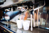 A close-up of an espresso machine brewing coffee into two white cups against a backdrop reminiscent of the Coffee 10 Wall Mural. The metallic spout drips rich espresso, with a black handle extending towards the viewer, as light highlights the shiny surface and drip tray below.