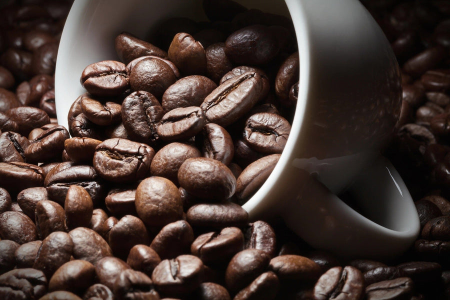 A modern coffee shop with wooden tables, stools, small potted plants, pendant lights, and large windows features the "Brewed to Perfection Wall Mural" depicting coffee beans spilling from a white cup as its centerpiece.