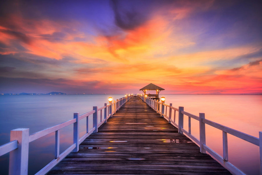A modern living-dining room with wooden floors and stylish furniture features the Bridge Made of Wood Wall Mural as its centerpiece, depicting a sunset pier. Warm lighting and vibrant skies complete the cozy, inviting atmosphere.