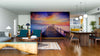 A modern living-dining room with wooden floors and stylish furniture features the Bridge Made of Wood Wall Mural as its centerpiece, depicting a sunset pier. Warm lighting and vibrant skies complete the cozy, inviting atmosphere.