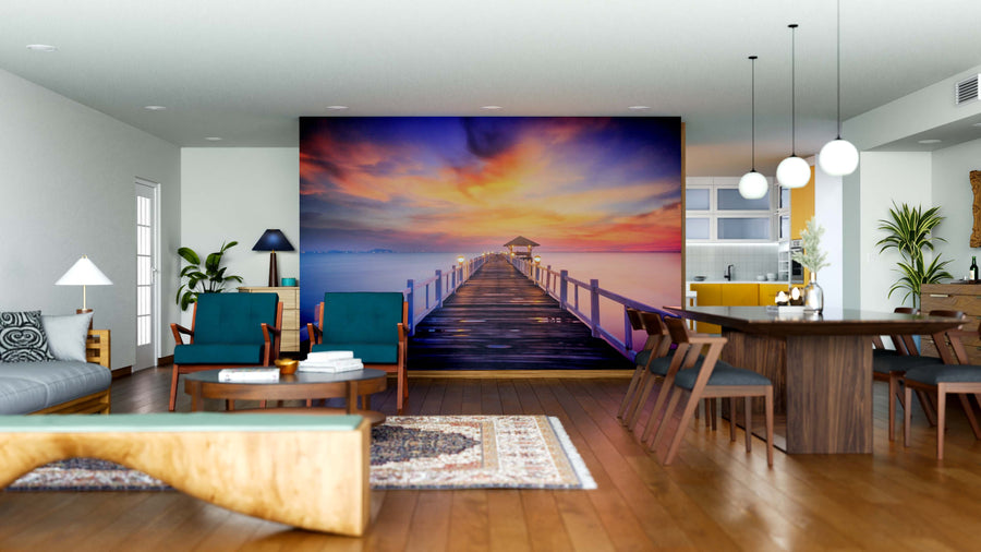 A modern living-dining room with wooden floors and stylish furniture features the Bridge Made of Wood Wall Mural as its centerpiece, depicting a sunset pier. Warm lighting and vibrant skies complete the cozy, inviting atmosphere.