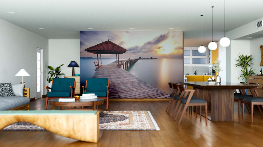 A modern open living-dining space features wooden furnishings, blue chairs, a patterned rug, and the "Bridge Over the Sea Wall Mural" on the back wall, adding a tranquil, scenic sunset atmosphere to the room.