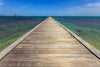 A wooden pier extends into the clear ocean, resembling the "Bridge to the Sea Wall Mural" by Deposit Photo. The water blends turquoise and deep blue with small islands in sight, giving an endless perspective.