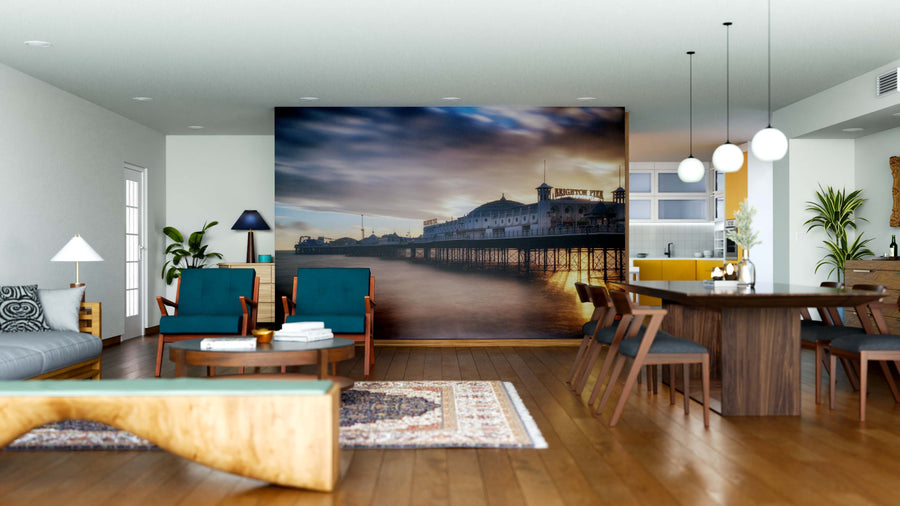 A modern living-dining area with wooden floors, teal armchairs, pendant lights, a dark dining table, patterned rug, and an open layout filled with natural light features the Brighton Pier Wall Mural as a striking focal point.