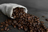 The Burlap Bag of Beans Wall Mural features dark roasted coffee beans spilling from a burlap sack onto rustic wood, creating rich texture and warmth for any space with easy-to-install, removable wallpaper.