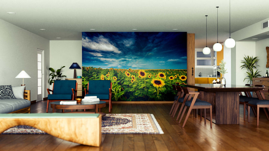 A modern living-dining room features wooden floors, mid-century furniture, and the Calm Before the Bloom Wall Mural—a vibrant sunflower field under a dramatic sky—that brings color and nature to the sleek space. Round lights hang above the dining table.