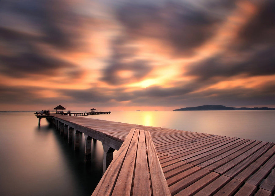 A modern living and dining area features wooden floors, stylish furniture, pendant lights, and the Calm Ocean Walkway Wall Mural—a striking peel and stick mural of a dock stretching into a tranquil lake at sunset with vibrant clouds.