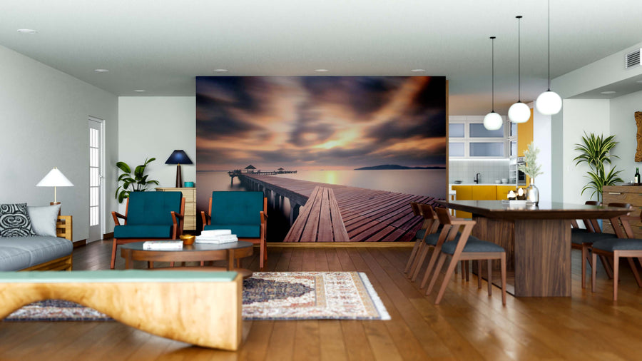 A modern living and dining area features wooden floors, stylish furniture, pendant lights, and the Calm Ocean Walkway Wall Mural—a striking peel and stick mural of a dock stretching into a tranquil lake at sunset with vibrant clouds.