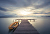 The "Calm Waters at Sunset Wall Mural" by Deposit Photo showcases a tranquil landscape with a wooden pier extending into a calm lake under a cloudy sky. A blurred orange boat adds dynamic movement to the serene scene as the setting sun casts a soft glow on the horizon.