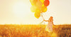 A young girl in a white dress with yellow and orange balloons stands in a sunlit field of tall grass, evoking the dreamy essence of the "Inspirational 2 Wall Mural," as her joyful smile glows warmly in the sunset's embrace, highlighting the summer atmosphere.