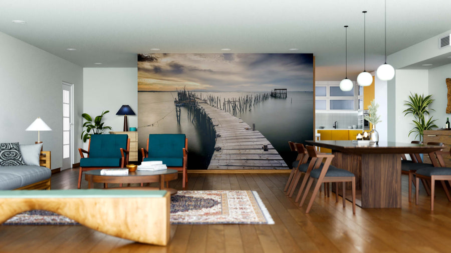 A modern, open living-dining area with wood floors, teal chairs, a Carrasqueira Old Wooden Pier Wall Mural, wooden dining table with dark chairs, pendant lights, indoor plants, and a kitchen featuring yellow accents in the background.
