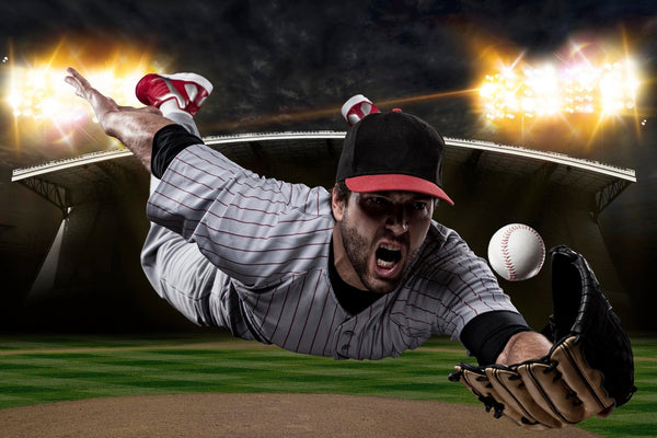 The Catching Baseball Wall Mural shows a player in a gray pinstripe uniform and black cap diving on a night field, arm outstretched with glove to catch the ball. Bright stadium lights illuminate the dramatic canvas-textured scene.