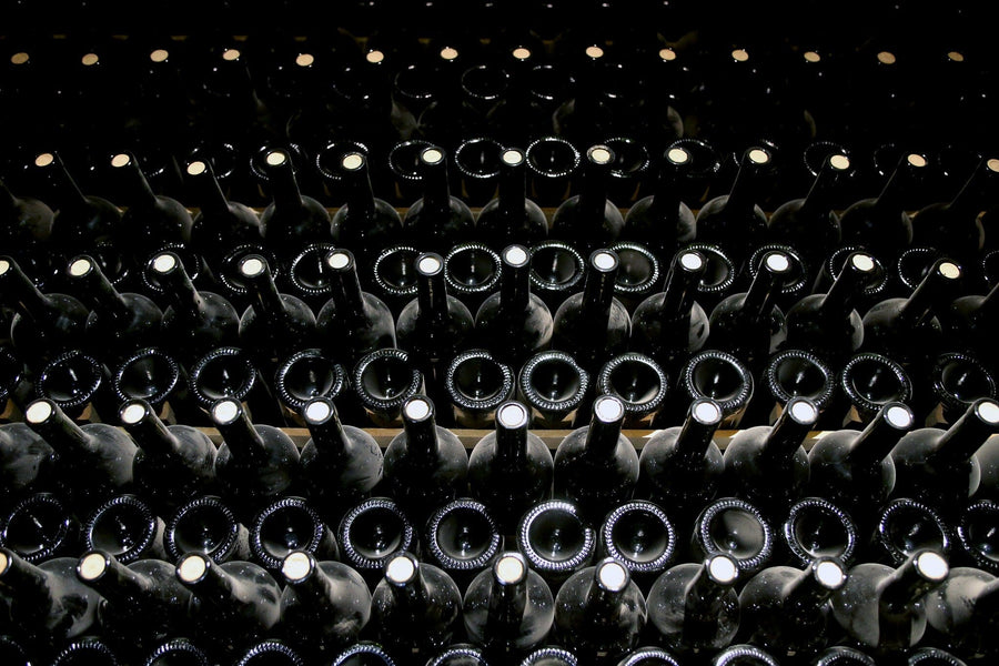 Modern cafe interior features wooden tables, black chairs, small potted plants, and the Cellar Stacked Bottles Wall Mural showing rows of wine bottles. Pendant lights hang above, with shelves and a coffee machine on the right.