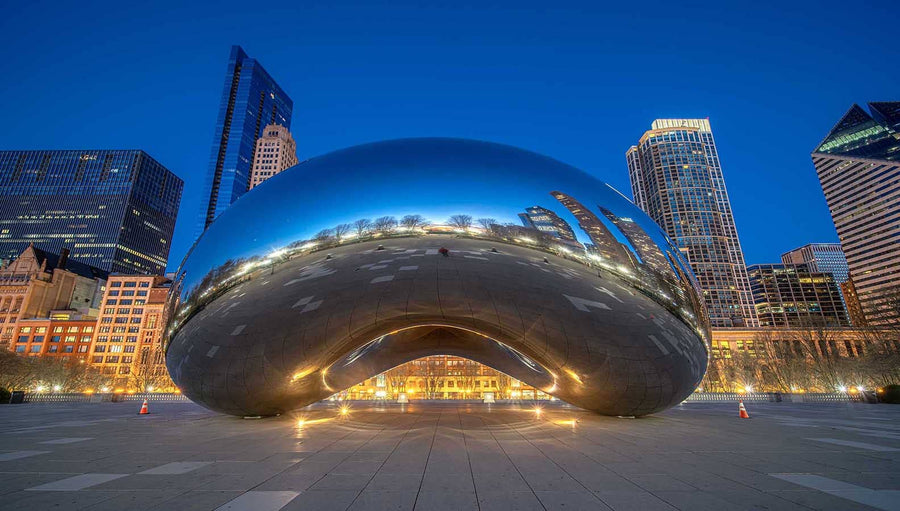 A contemporary bedroom displays a "Chicago Bean at Dusk" wall mural reflecting city lights, featuring a green upholstered bed with gray bedding, wooden nightstands, built-in shelving, and a large window to the left that fills the room with natural light.