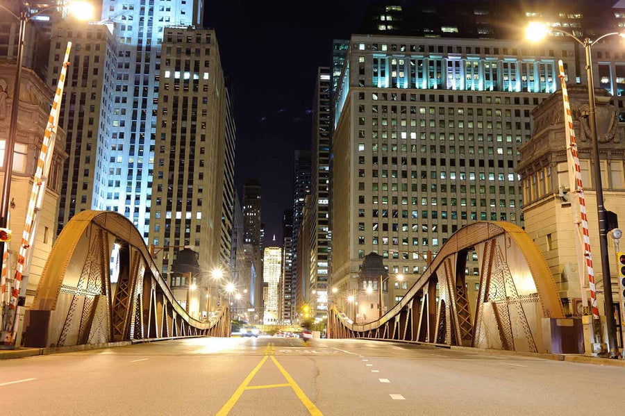 The Chicago Downtown Bridge at Night Wall Mural showcases a well-lit urban scene with an arching bridge leading to a cityscape. The mural features tall buildings, curved beams, overhead lights, red and white barriers, and empty streets—perfect for a stunning custom-printed mural.