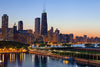 The Chicago Dusk at Belmont Harbor Wall Mural portrays the city's skyline with lit skyscrapers like Willis Tower, reflecting on tranquil water. The orange and blue evening sky, trees, and a boat add serenity to the vibrant city in this urban-themed peel and stick mural.