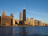 A custom printed mural titled "Chicago Morning On Hancock Building Wall Mural" features a stunning skyline view of Chicago, showcasing prominent skyscrapers such as the dark Willis Tower and the beige John Hancock Center. The buildings beautifully reflect sunlight under a clear blue sky, while Lake Michigan's calm surface contributes to a serene urban and waterfront landscape.