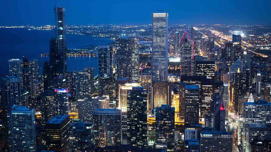 A modern conference room with black chairs and a long wooden table features the "Chicago Skyscrapers Aerial View at Night Wall Mural"; a blue rug lies beneath, and glass walls reveal another office area.