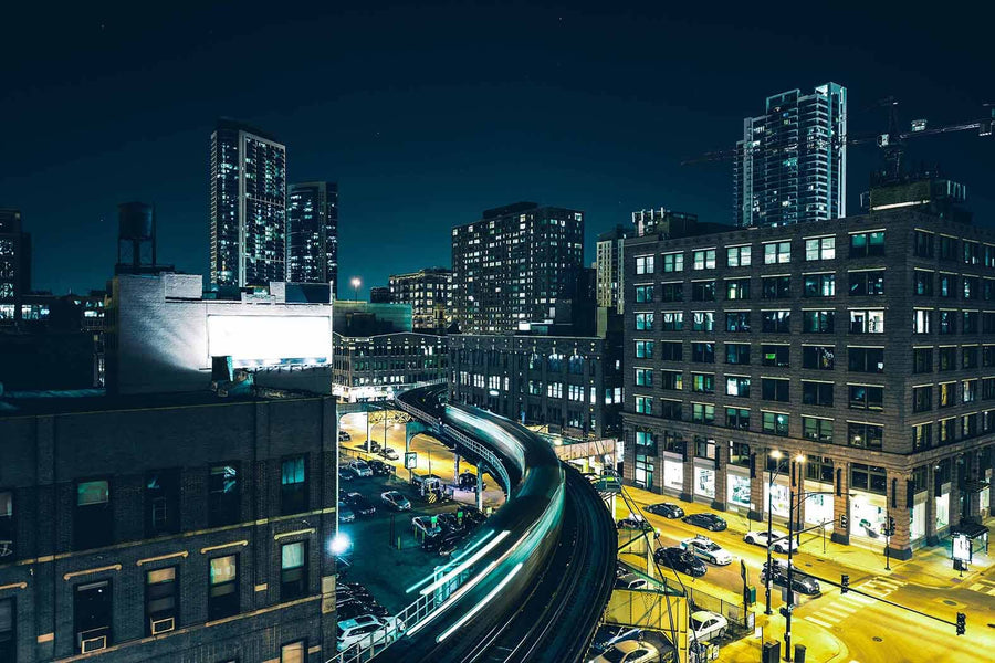 A modern conference room displays the "Chicago Speeding Train At Night" wall mural with its vibrant cityscape featuring illuminated buildings and busy streets. It includes a long white table, black chairs, wooden walls, and three gray pendant lights overhead.