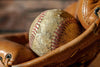 The Classic Leather and Stitch Wall Mural features a close-up of a weathered baseball with red stitching in a well-used brown glove, creating nostalgic charm for any room with its soft, blurred background. Peel and stick for easy decor.