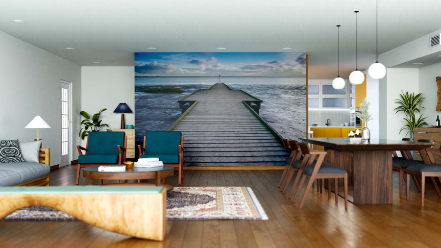 A modern, open-plan living and dining space with wood floors, teal and wood furniture, pendant lights, and the Coastal Solitude Wall Mural featuring a boardwalk over the ocean under a cloudy sky creates a serene, spacious vibe.