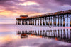 Under a dramatic sunset sky in shades of purple, orange, and pink, the Cocoa Beach Wall Mural depicts a long wooden pier extending into the ocean. Serene reflections dance on wet sand as a solitary bird stands by the water's edge while waves lap against the pier.