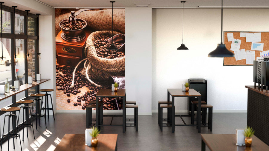 A modern café features wooden tables and chairs, bar seating by large windows, and the Bean to Brew Wall Mural showing coffee beans, a grinder, and a burlap sack. Pendant lights hang above, with small plants on each table.