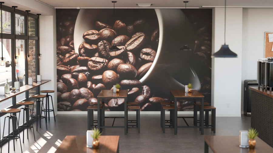 A modern coffee shop with wooden tables, stools, small potted plants, pendant lights, and large windows features the "Brewed to Perfection Wall Mural" depicting coffee beans spilling from a white cup as its centerpiece.