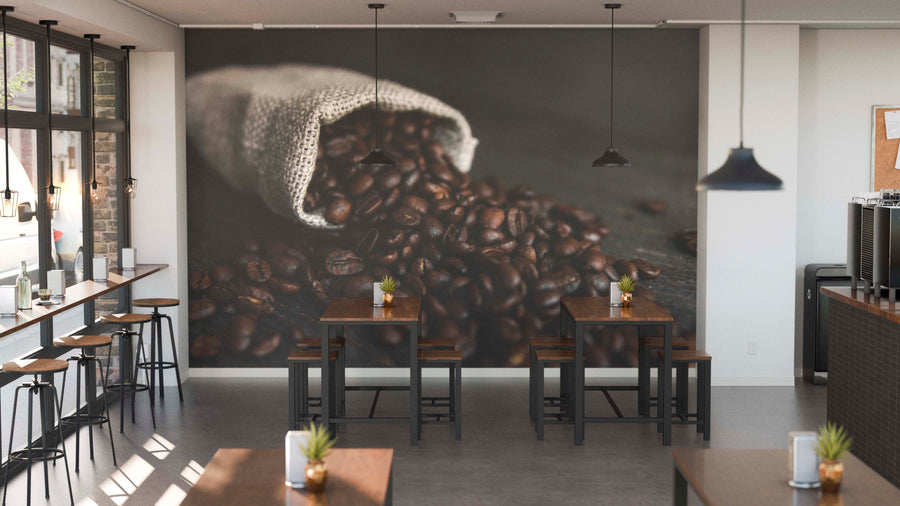Modern coffee shop with wooden tables, black chairs, and potted succulents. The Burlap Bag of Beans Wall Mural shows roasted beans spilling from a sack. Industrial pendant lights hang above, and tall windows fill the space with natural light.