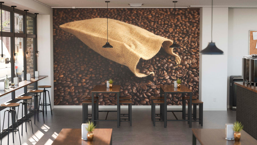A modern coffee shop features wooden tables, stools, potted plants, large windows, and a Burlap Sack of Coffee Beans Wall Mural. Black pendant lamps hang overhead, completing the inviting space.