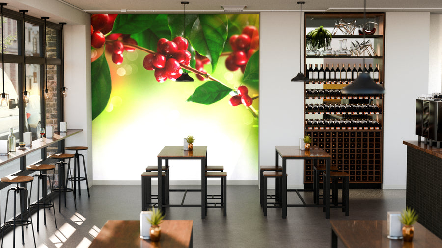 A modern café features wooden tables, black chairs, and natural light. One wall displays the vibrant Coffee Cherry Branch Wall Mural, while shelves with bottles and plants line another. Each table is decorated with a small plant.