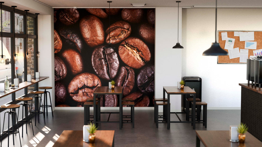 A modern coffee shop features wooden tables and stools, potted plants, a corkboard, large windows, and a Close-Up of Roasted Coffee Beans Wall Mural. Soft lighting adds to the inviting atmosphere.