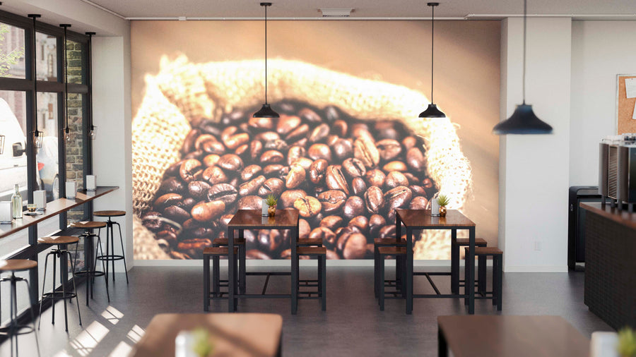 A modern café interior with wooden tables, stools, pendant lights, and sunlit windows features the Bag of Coffee Beans Wall Mural on the back wall. Small plants and tableware decorate the tables for a fresh touch.