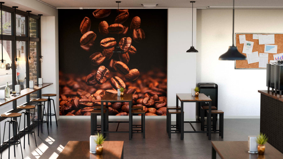 A modern café features wooden tables and stools, large left-side windows, and the Coffee Beans Falling Wall Mural as a focal point. Pendant lights hang above, with small plants on tables next to a bulletin board on the right wall.