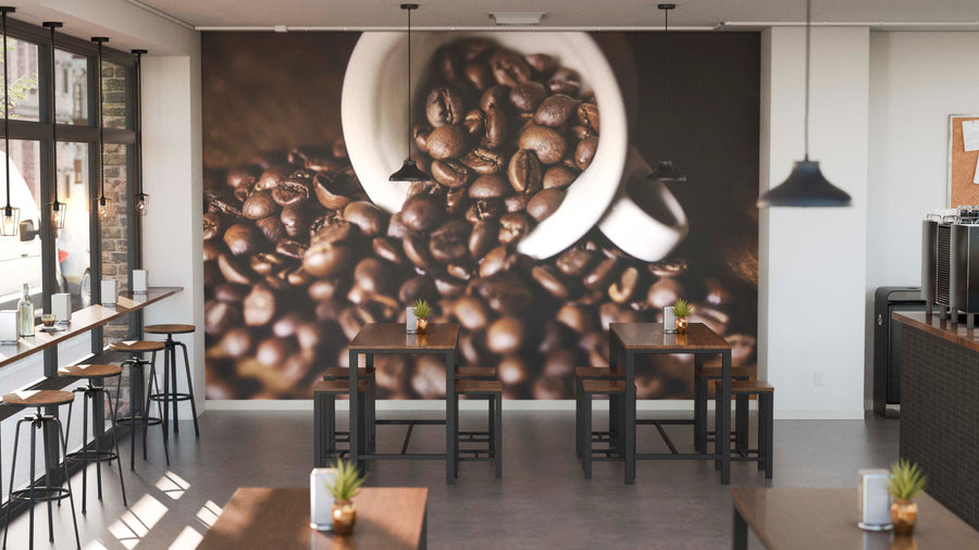 A modern coffee shop with wooden tables, stools, potted plants, and hanging lights features the Coffee Beans Spilling Wall Mural—a custom print showing a white cup spilling dark roasted beans. Large windows on the left fill the space with natural light.