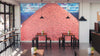 A modern café with wooden tables and chairs features the Bounty of Beans Wall Mural on its back wall. Pendant lights hang from the ceiling, sunlight streams in, and small plants adorn the tables.