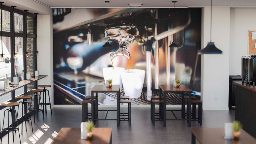 A modern coffee shop interior features wooden tables and stools, potted succulents, black pendant lights, and the Brewed Espresso Wall Mural showcasing a vivid close-up of espresso pouring into a white cup.