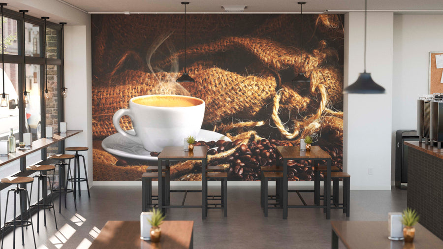 A modern café interior with wooden tables, potted plants, and pendant lights. The back wall displays the Coffee Farm in Colombia Wall Mural, showing a coffee cup on a saucer among beans and burlap sacks. Sunlight streams through tall windows.