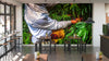 A café interior with modern wooden tables and chairs features the Coffee Farmer Wall Mural, depicting a person in a blue shirt harvesting red coffee cherries into a yellow basket amid lush greenery, sunlight streaming through tall windows for added warmth.