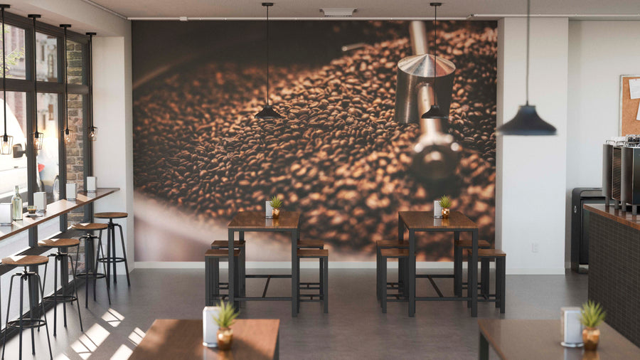 A modern café with wooden tables, stools, plant centerpieces, and big left-side windows features the Coffee Roast Wall Mural of coffee beans on the back wall. Pendant lights overhead enhance the cozy and inviting atmosphere.