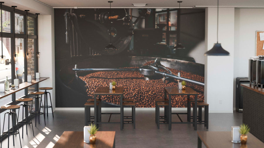 A modern café with wooden tables, stools, potted plants, and large windows features the Coffee Roasting Process Wall Mural. Pendant lights hang overhead, creating a warm and inviting atmosphere.