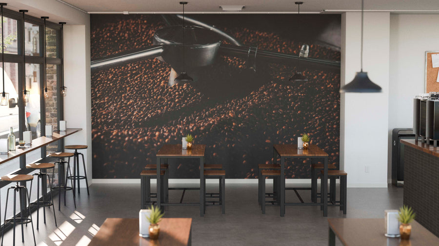 A modern café interior with wooden tables, high stools, potted plants, and the “Coffee in the Making Wall Mural” as a feature wall. Pendant lights hang overhead while sunlight streams through tall windows on the left.