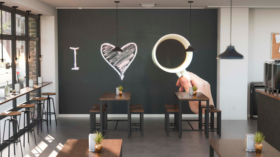 A modern café with wooden tables and potted plants features the Coffee is Love Wall Mural—a large chalk-style “I ♥️” and hand with a coffee cup—illuminated by daylight from big windows in the empty space.