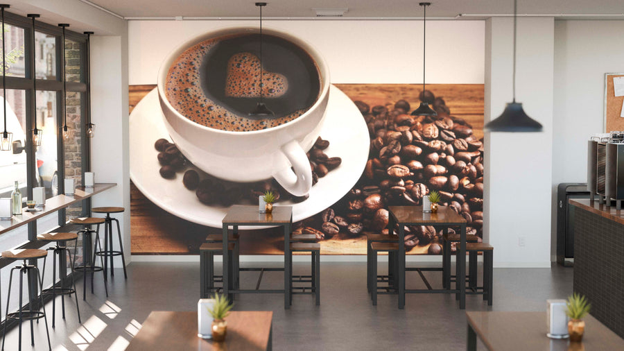 A bright, modern coffee shop with wooden tables and potted plants features the "Cup Full of Coffee Beans Wall Mural"—a peel and stick mural showcasing a coffee cup with heart-shaped foam and beans, adding warmth and charm to the space.