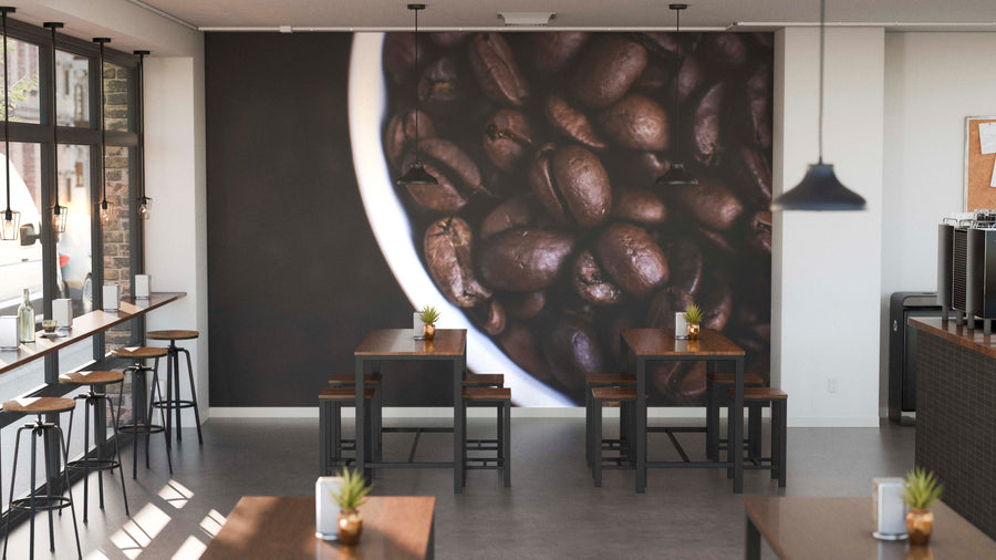 Modern coffee shop interior featuring wooden tables, stools, potted plants, and a striking Cup of Coffee Beans Wall Mural. Pendant lights and large windows fill the space with natural light.