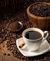 A Coffee Cup with Cinnamon Stick Wall Mural features a steaming black coffee on a white saucer, surrounded by coffee beans and ground coffee in bowls, with a cozy café scene depicted on the rustic wooden surface.