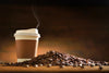 A steaming takeaway coffee cup with a brown sleeve sits next to scattered beans on wood, set against a warm, blurred gradient backdrop. The scene evokes a cozy atmosphere similar to those featured in the Coffee 13 Wall Mural