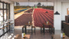 A modern café features wooden tables, stools, potted plants, and a back wall adorned with the Drying Yard Wall Mural—coffee cherries drying outdoors beneath a vibrant blue sky with distant trees.